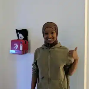 Vrouw met duim omhoog naast Philips HeartStart HS1 AED in transparante wandhouder.