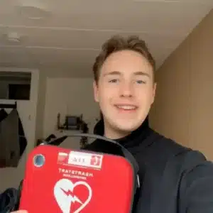 Jonge man toont glimlachend een Philips AED in rode draagtas in een binnenruimte.