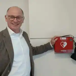 Man in pak wijst glimlachend naar een Philips AED in rode wandhouder aan een witte muur.