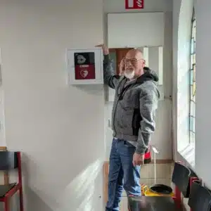 Man wijst trots naar nieuw geplaatste AED in wandkast aan de muur.