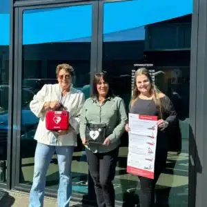 Drie vrouwen poseren buiten met een Philips AED, opbergtas en reanimatie-instructieposter.