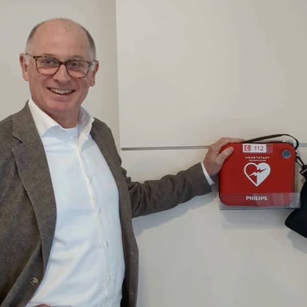 Man in pak wijst glimlachend naar een Philips AED in rode wandhouder aan een witte muur.