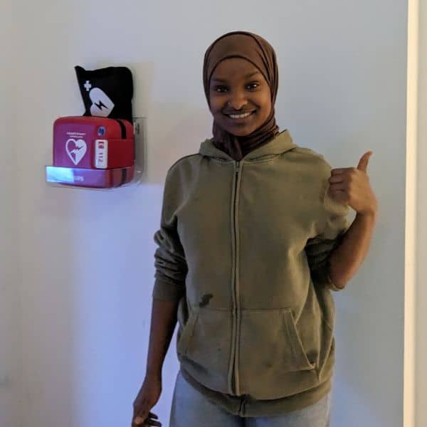 Vrouw met duim omhoog naast Philips HeartStart HS1 AED in transparante wandhouder.