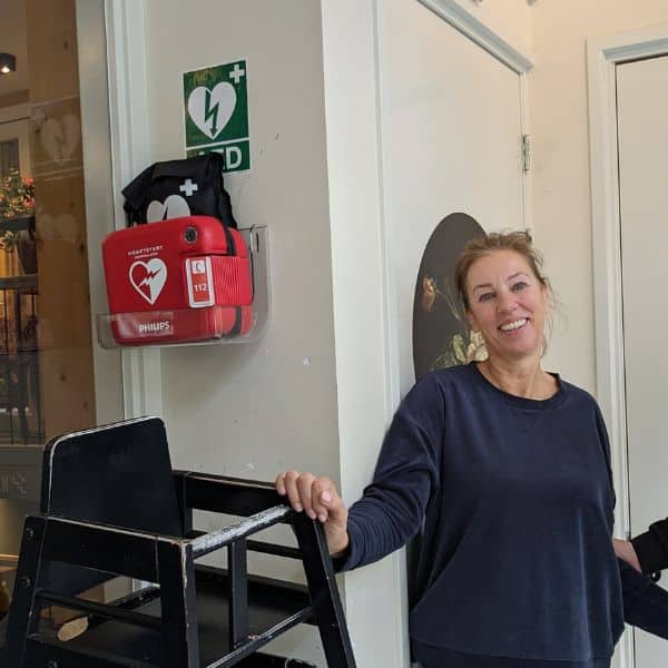 Vrouw staat binnen naast een wandgemonteerde Philips AED in houder met AED-bord erboven.