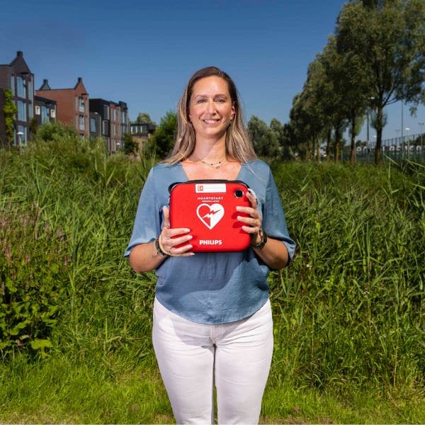 Vrouw houdt een Philips HeartStart HS1 AED vast in een groene woonwijkomgeving.