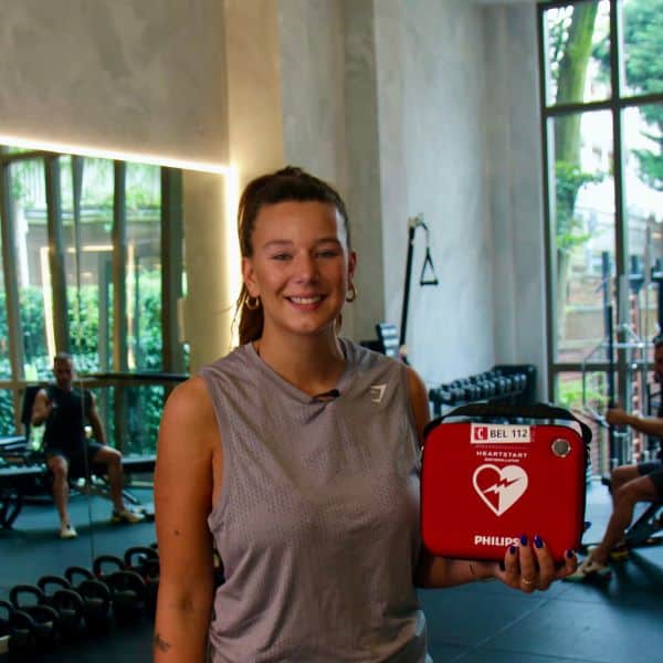 Vrouw in sportoutfit houdt een Philips HeartStart HS1 AED vast in een fitnessruimte.