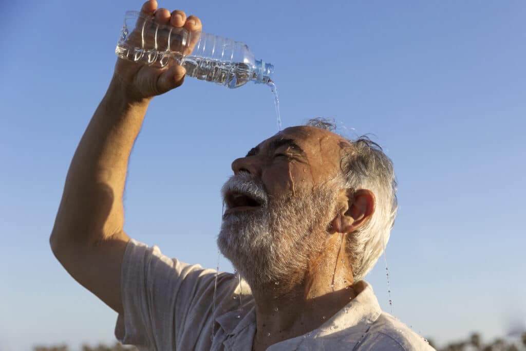 Oudere man schenkt een flesje water leeg over zijn voorhoofd om af te koelen tijdens warm weer.